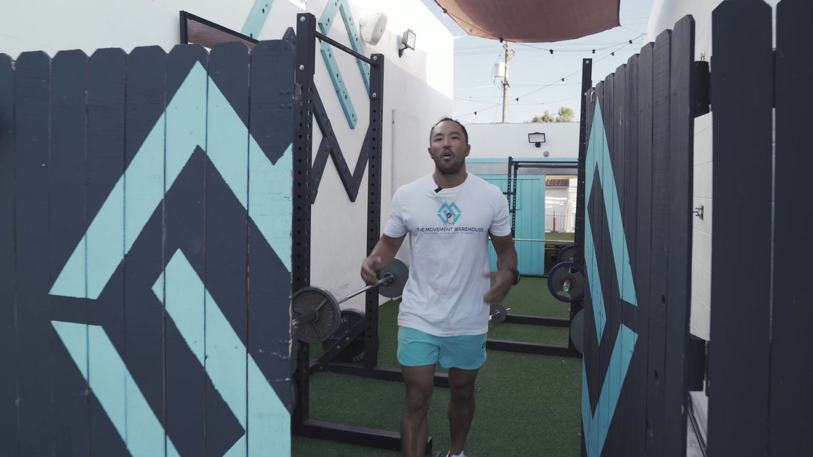 A coach holding a barbell between the black and sky-blue fence panels at MVMT